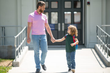 A father and son walk out of a school together holding hands.